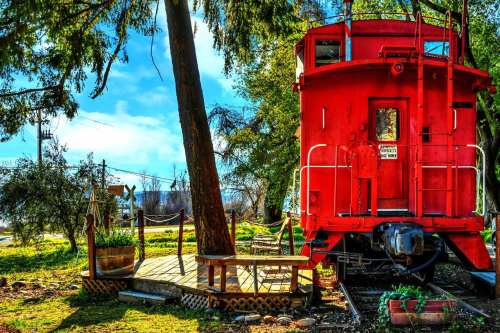 A bright red caboose sits among trees, with a wooden deck and potted plants in a sunny outdoor setting.