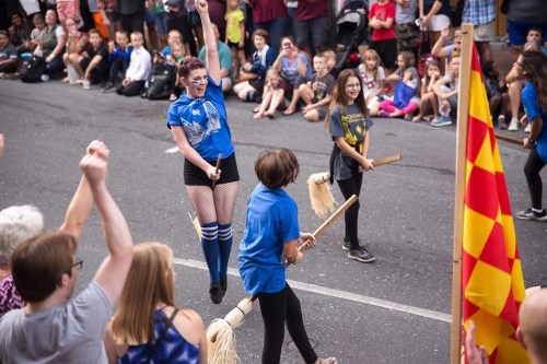 A lively street performance with dancers in blue shirts and broomsticks, entertaining a cheering crowd.