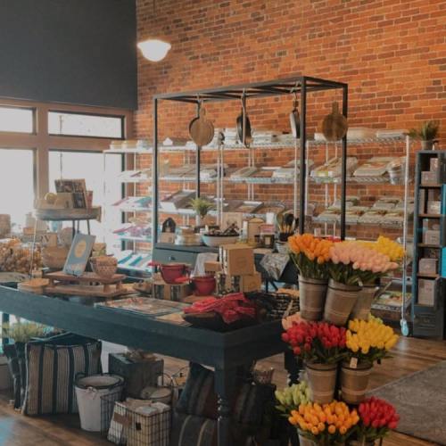 A cozy shop interior with brick walls, shelves of goods, and vibrant flower arrangements on display.