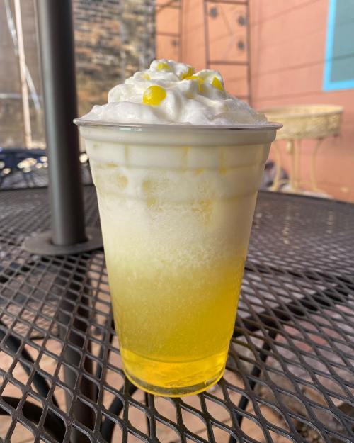 A frosty drink in a clear cup, featuring a yellow gradient, topped with whipped cream and yellow candy pieces.