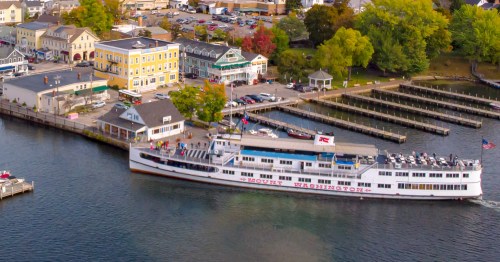 Aerial view of a large boat docked near colorful buildings and a marina, surrounded by trees and parked cars.
