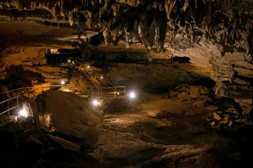 Dimly lit cave interior with stalactites, rocky terrain, and a metal walkway illuminated by soft lights.
