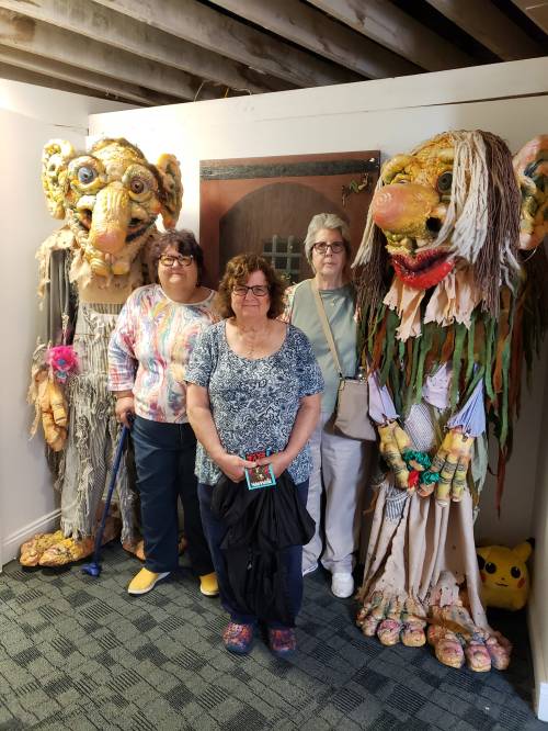 Three women pose in front of large, colorful puppet-like figures in a whimsical indoor setting.