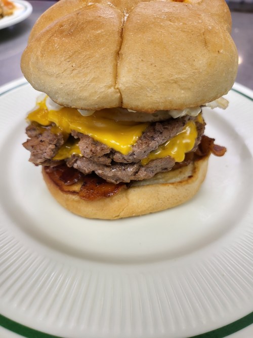 A stacked burger with two beef patties, melted cheese, bacon, and a soft bun on a white plate.