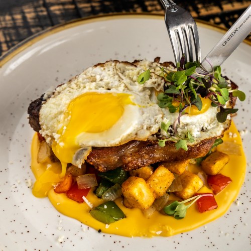A plate of steak topped with a fried egg, served over colorful diced vegetables and cheese sauce.