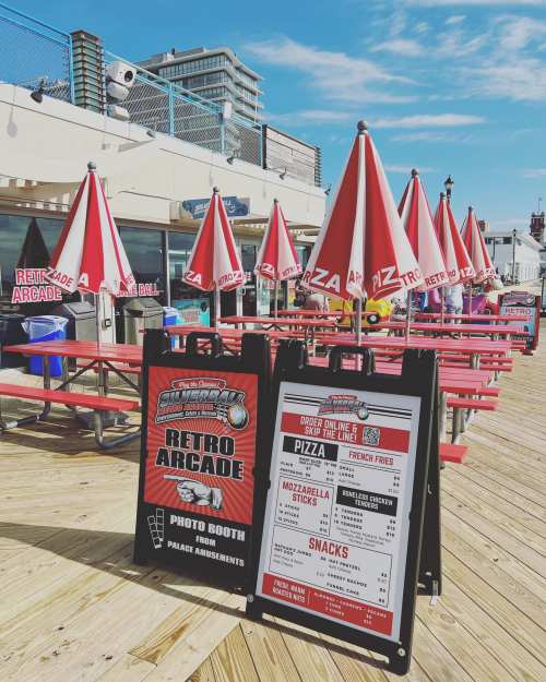 A vibrant boardwalk scene featuring a retro arcade, picnic tables, umbrellas, and a menu board for pizza and snacks.