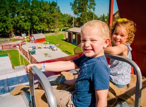 Two smiling children sit on a ride, enjoying a sunny day at an amusement park with a scenic view behind them.