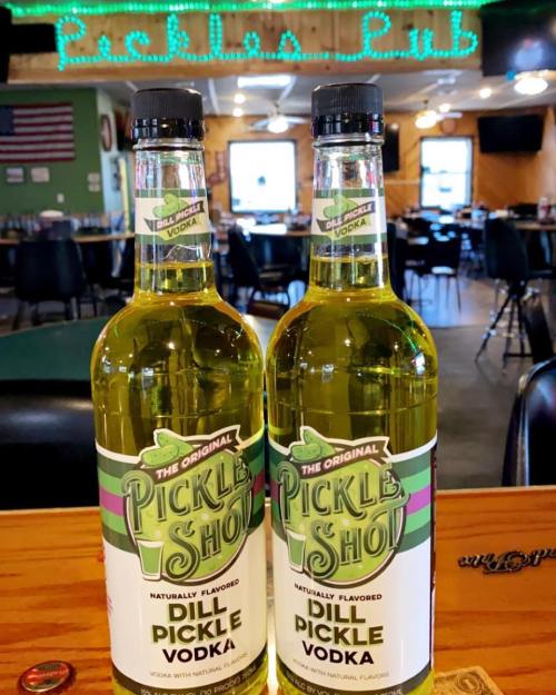 Two bottles of Dill Pickle Vodka on a wooden bar, with a neon "Pickles Pub" sign in the background.