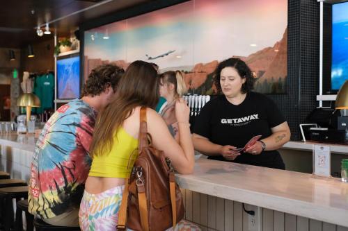 A bartender assists two customers at a bar with a colorful backdrop of mountains and a plane in the sky.