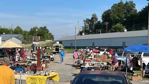 A bustling outdoor flea market with various stalls, people browsing, and cars parked nearby under a clear blue sky.