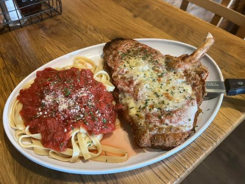 A plate of breaded pork chop topped with cheese, served with fettuccine pasta and marinara sauce, garnished with herbs.