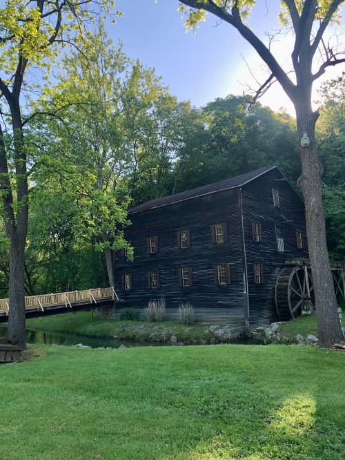 A historic black wooden mill beside a pond, surrounded by lush green trees and a wooden bridge.