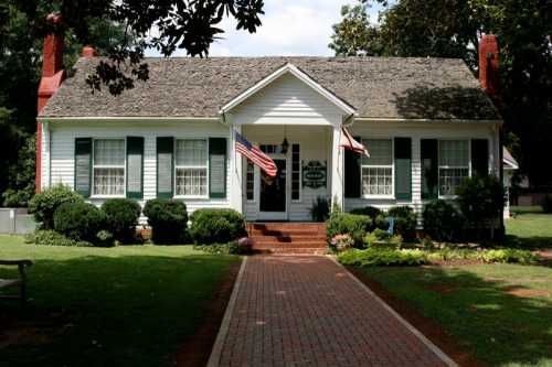 A charming white house with green shutters, a brick path, and American flags in a landscaped yard.