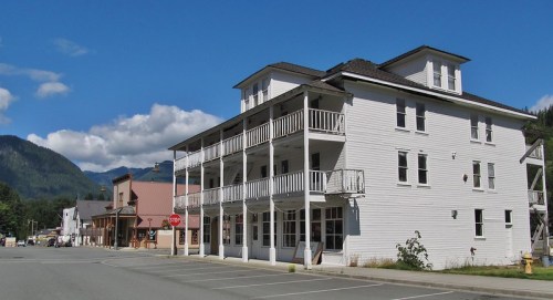 A white, two-story building with a balcony, set against a backdrop of mountains and a clear blue sky.