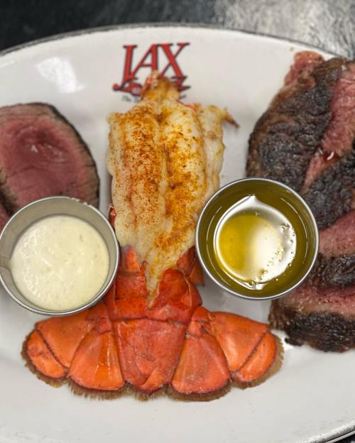 A plate featuring lobster tail with seasoning, steak, and two dipping sauces.