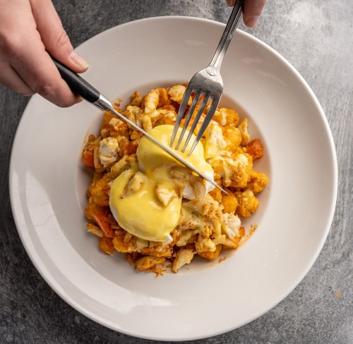 A hand holding a fork and knife cuts into a plate of eggs Benedict on a bed of cauliflower and crab.