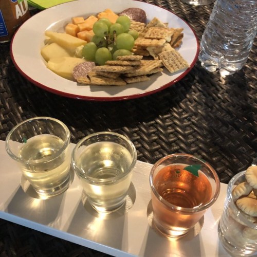 A platter of cheese, crackers, and fruit alongside four shot glasses with different drinks on a table.