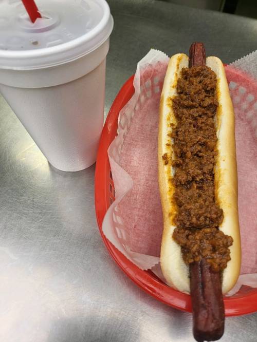 A hot dog topped with chili in a red basket, next to a cup of drink.