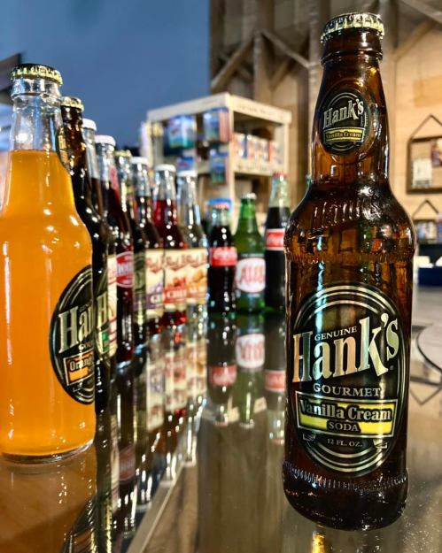 A close-up of a Hank's Vanilla Cream soda bottle in front of a variety of colorful soda bottles on a table.