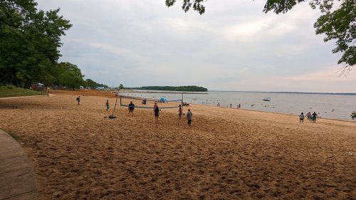 A sandy beach with people playing volleyball, swimming, and relaxing by the water under a cloudy sky.