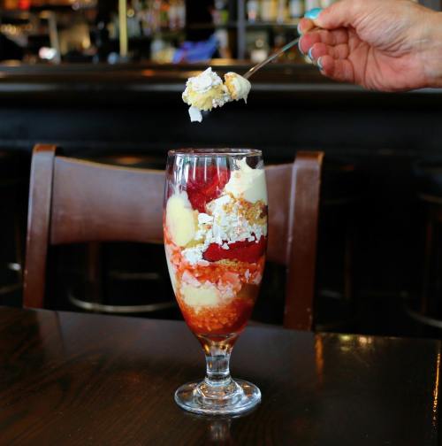 A glass dessert parfait layered with strawberries, cream, and cake, with a spoonful being lifted from the top.