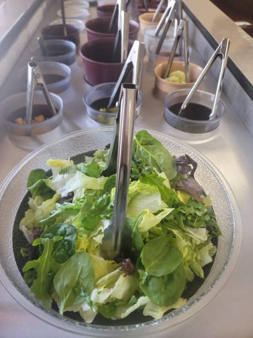 A bowl of fresh mixed greens with tongs, surrounded by various small containers of dressings and toppings.