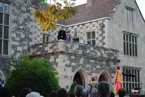 A theatrical performance outside a stone building, featuring actors on a balcony and others on the ground.