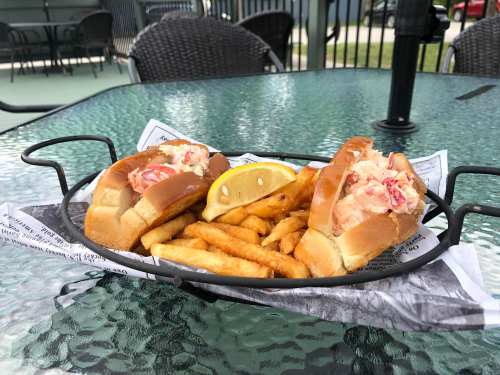 Two lobster rolls with a side of fries and a lemon wedge, served on a black wire basket on a glass table.