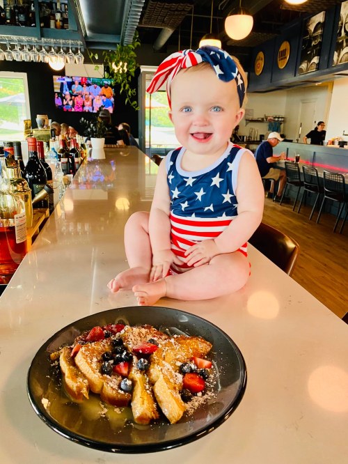 A smiling baby in a patriotic swimsuit sits on a bar counter next to a plate of French toast topped with berries.