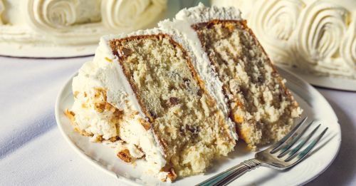 A slice of layered cake with white frosting and nuts, served on a plate with a fork beside it.