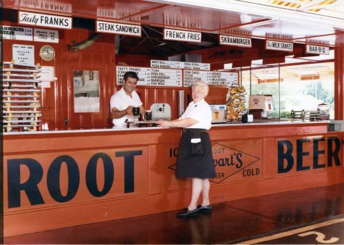 A vintage diner counter with a man serving food and a woman in a uniform, featuring a menu of classic snacks and drinks.