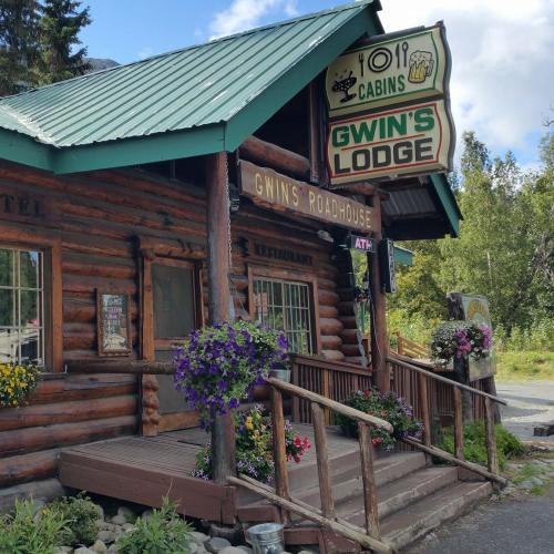 A rustic log cabin lodge with a green roof, featuring a sign for Gwin's Roadhouse and colorful flower pots.