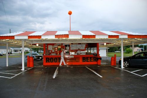 A retro-style root beer stand with a striped roof, outdoor seating, and cars parked nearby under a cloudy sky.