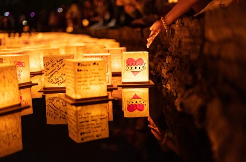 Lanterns with handwritten messages float on water, surrounded by people reaching out to them in a serene evening setting.