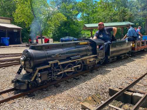 A steam locomotive on tracks with a man in a cap operating it, surrounded by trees and a small station.