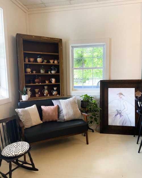 Cozy interior featuring a wooden shelf with pottery, a black sofa with cushions, and a framed artwork by a window.