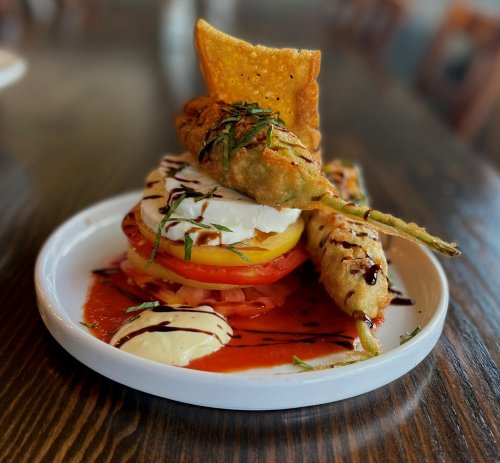 A colorful plate featuring layered vegetables, mozzarella, and crispy fried items drizzled with sauce.