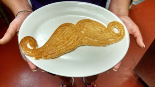A person holds a plate with a pancake shaped like a mustache.