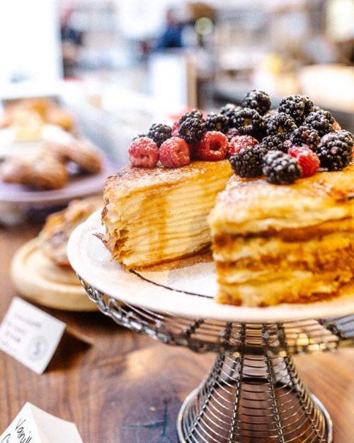 A layered cake topped with fresh berries, displayed on a cake stand, with other pastries in the background.