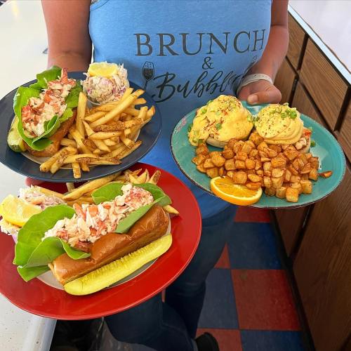 A person holds two plates of food: lobster rolls with fries and eggs Benedict with home fries, wearing a "Brunch & Bubbles" shirt.