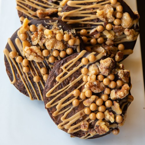 Three chocolate cookies topped with caramel drizzle, crunchy nuts, and golden candy pearls on a white plate.