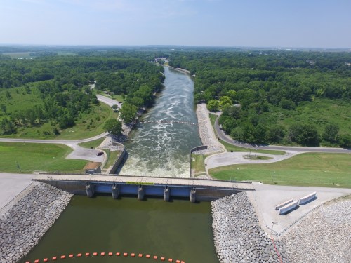 Aerial view of a river with a dam, surrounded by green trees and a winding road. Water flows over the dam.