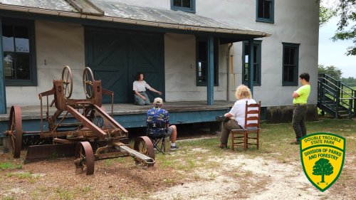A group of people sitting on a porch with a vintage tractor nearby, surrounded by greenery at Double Trouble State Park.