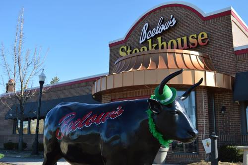 A black cow statue wearing a green hat stands in front of Beelow's Steakhouse, featuring a brick exterior and sign.