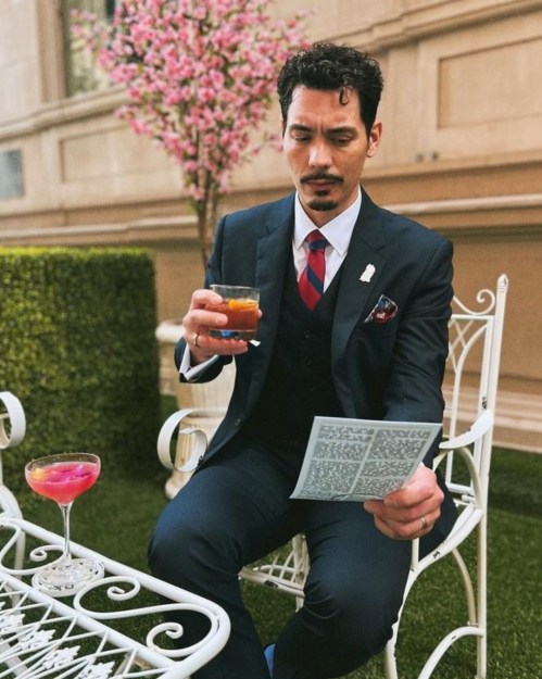 A man in a suit sits on a white chair, holding a drink and reading a menu, with pink flowers in the background.
