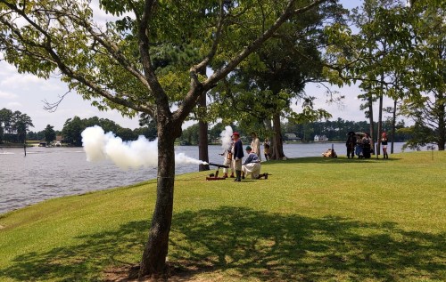 A group of people near a lake, with smoke rising from a cannon demonstration under a tree on a sunny day.