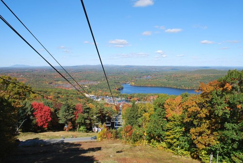 A scenic view from a ski lift, showcasing colorful autumn trees and a lake under a clear blue sky.