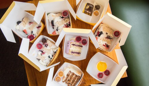 A variety of desserts in takeout boxes, including cakes and pastries, arranged on a wooden table.