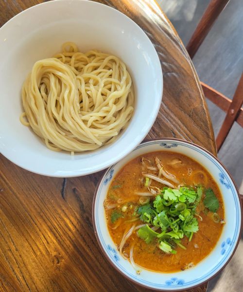 A bowl of rich, orange soup topped with herbs, alongside a separate bowl of plain noodles on a wooden table.