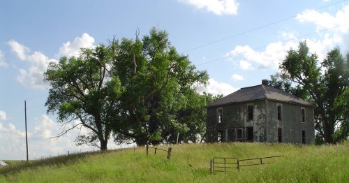 Abandoned two-story house surrounded by tall grass and trees under a blue sky with scattered clouds.